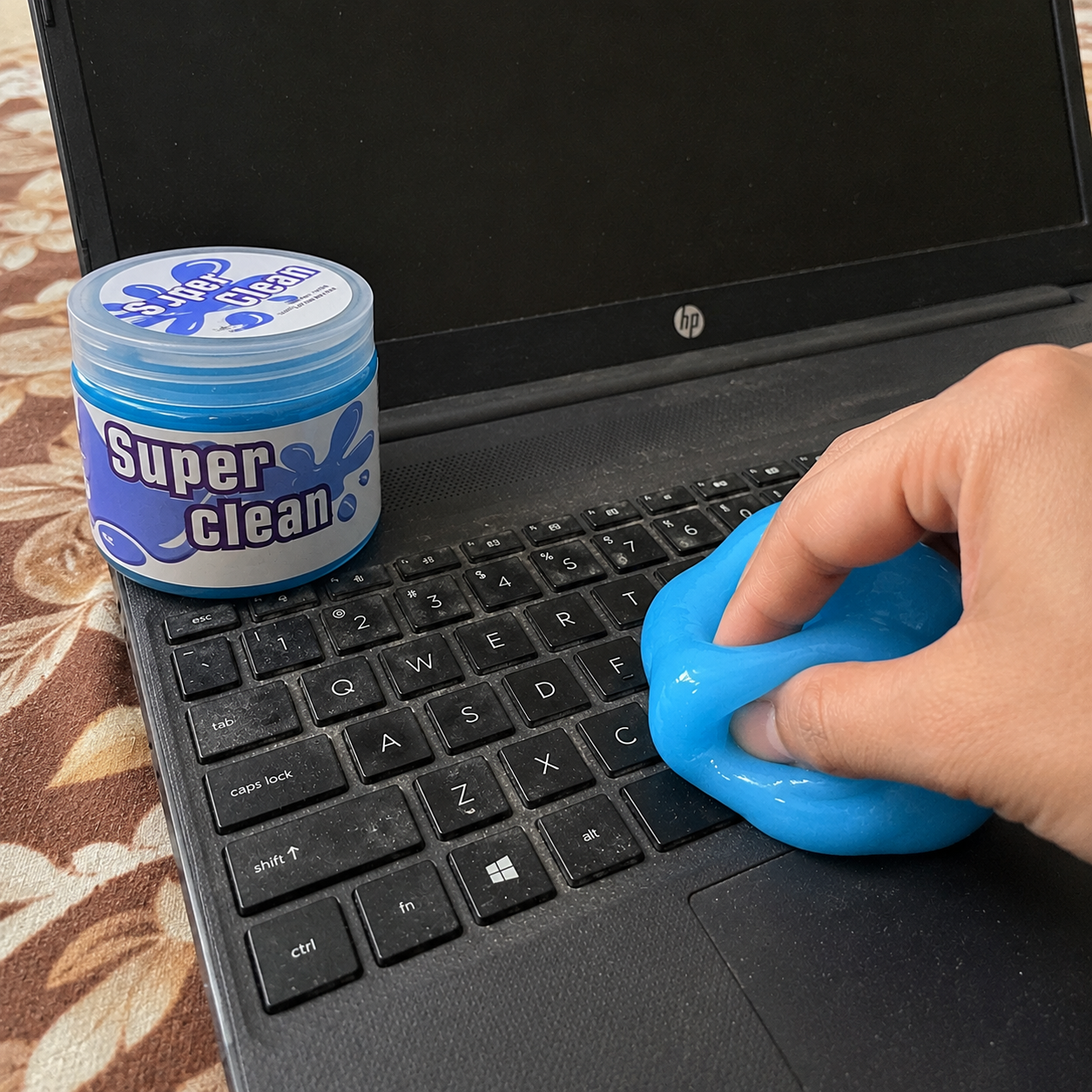 Person cleaning a laptop keyboard with a blue cleaning tool next to a container labeled 'Super Clean' on a patterned surface.